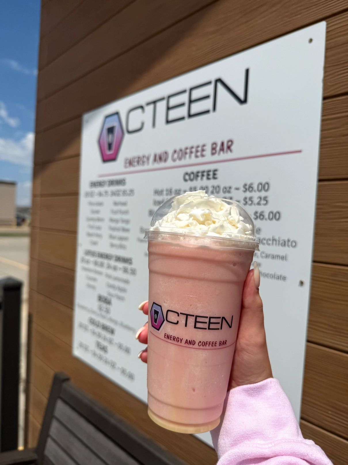 A hand holding a pink Octeen energy drink topped with whipped cream in front of the CTEEN menu board
